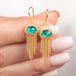 A pair of gold dangle earrings with oval emerald gemstones and cascading gold chain fringe, shown held in hand.