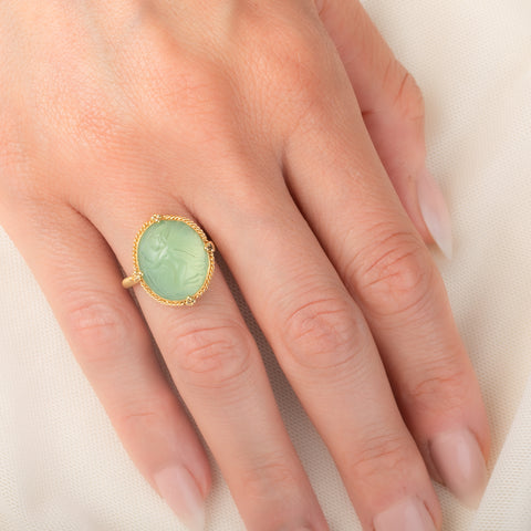 A gold ring featuring a prehnite with a fairy design carved in it, worn on a person's finger.