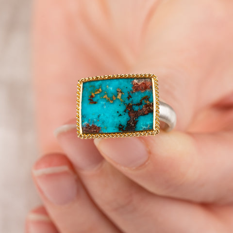 Close up of a hand holding a turquoise ring with gold bezel and silver band