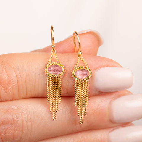 A pair of gold dangle earrings with oval pink rhodochrosite gemstones and cascading gold chain fringe, shown held in hand.
