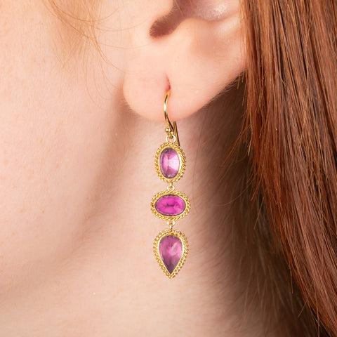 Gold earrings featuring a trio of vibrant red-pink rhodolite garnet gemstones, shown worn in a person's ear.