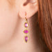 Gold earrings featuring a trio of vibrant red-pink rhodolite garnet gemstones, shown worn in a person's ear.