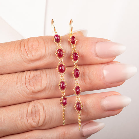 Gold earrings featuring a row of five ruby stones, shown held on a person's hand.