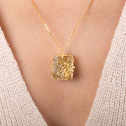 A gold pendant necklace featuring a rutilated quartz gemstone filled with fine metallic gold needles, worn on a person's neck.