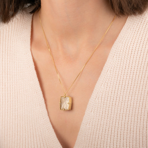 A gold pendant necklace featuring a rutilated quartz gemstone filled with fine metallic gold needles, worn on a person's neck.