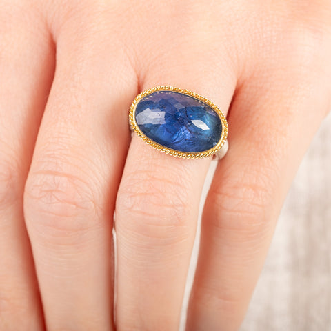 Close up of person wearing a handmade ring with blue tanzanite, gold bezel and sterling silver band on a white background