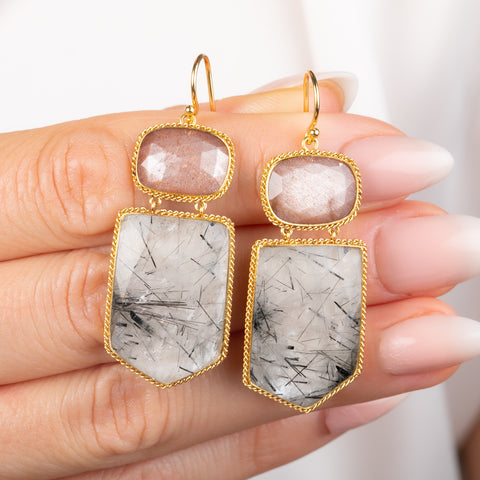 Gold earring featuring a tourmalated quartz and pink moonstone gemstone, displayed on a person's hand.