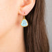 A close-up of a person's ear wearing a teardrop-shaped drop earring with a light blue turquoise stone and a gold bezel setting.
