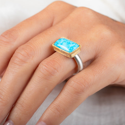 A gold ring featuring a rectangular turquoise stone set in a gold bezel with a sterling silver band on a person's finger