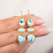 A pair of gold drop earrings with light blue turquoise stones, held between fingers against a soft, light beige background.