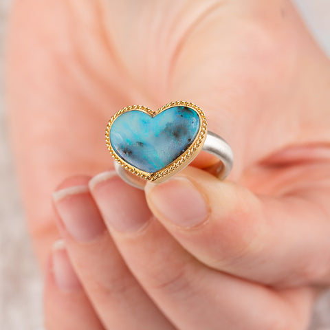 Hand holding a boulder opal heart ring with a gold bezel and silver band