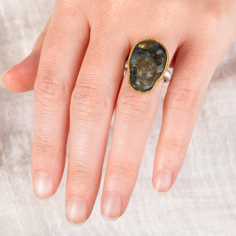 A hand wears a labradorite skull ring set in an 18k gold bezel with a sterling silver band