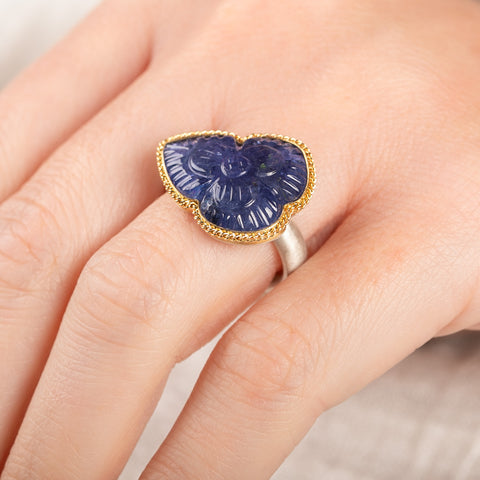 A person wears a handmade fine jewelry ring with carved tanzanite stone set in gold and complete with sterling silver band