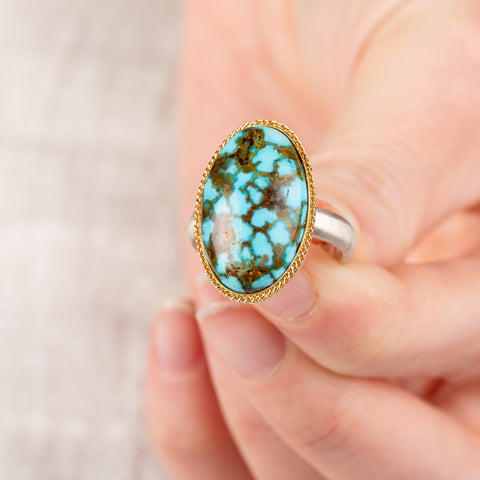 Close up of hand holding a turquoise ring with gold bezel and sterling silver band 
