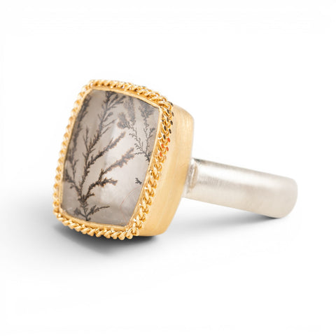 A gold and silver ring featuring a dendritic quartz stone with a detailed tree branch pattern inside, set against a clean white background.