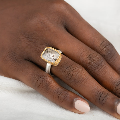 A gold and silver ring featuring a dendritic quartz stone with a detailed tree branch pattern inside, on a person's finger.