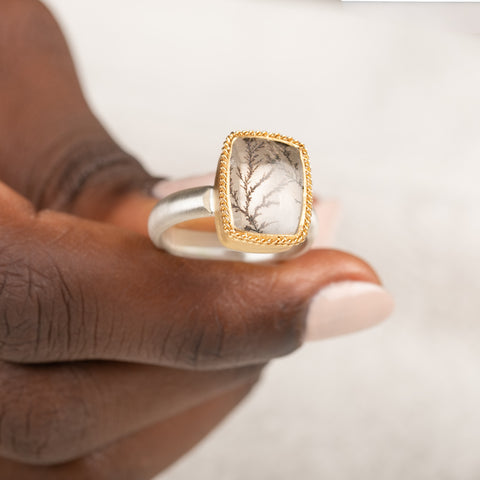 A gold and silver ring featuring a dendritic quartz stone with a detailed tree branch pattern inside, held in hand