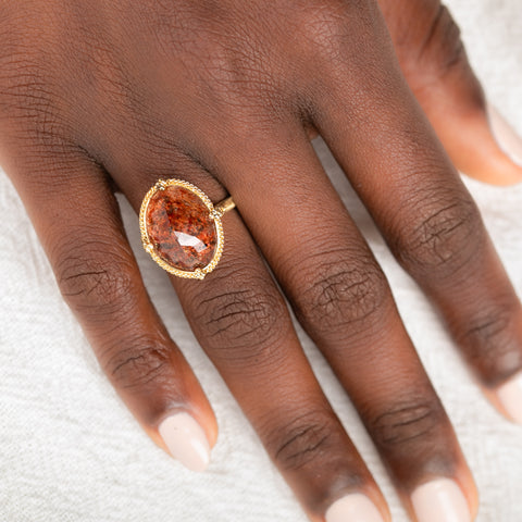 Gold ring featuring a sunstone gemstone on a person's finger against a neutral background