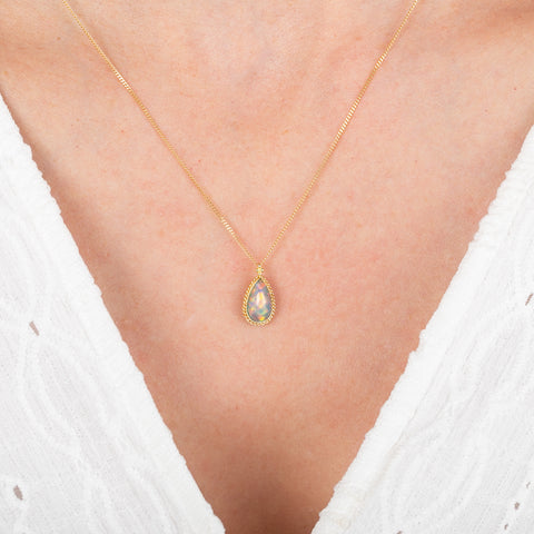 Gold necklace with an ethiopian opal teardrop-shaped pendant on a person wearing a white top.