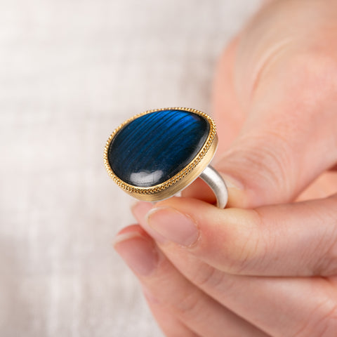 A hand holding a ring with labradorite stone set in a gold  bezel with a silver band