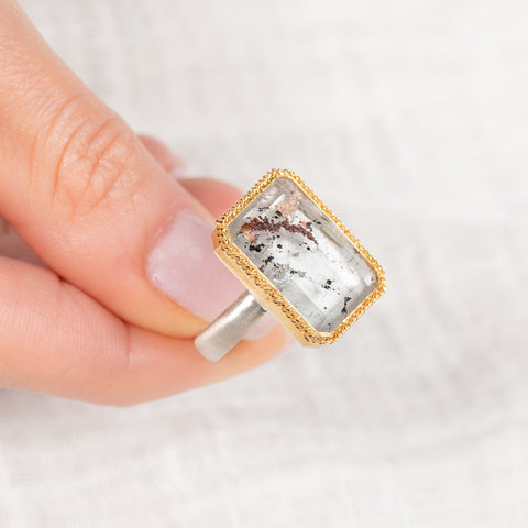 Gold and silver ring with a lodolite quartz stone held by a hand on a light background