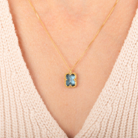 A gold necklace featuring a london blue topaz stone set in an 18k gold bezel, worn on a person's neck.