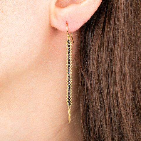 A pair of gold drop earrings with a row of black diamonds, worn in a person's ear.
