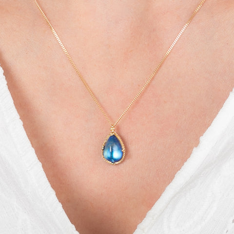 Gold necklace with a blue moonstone teardrop pendant on a person wearing a white top.