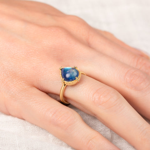 Gold ring with a blue moonstone on a hand against a neutral background