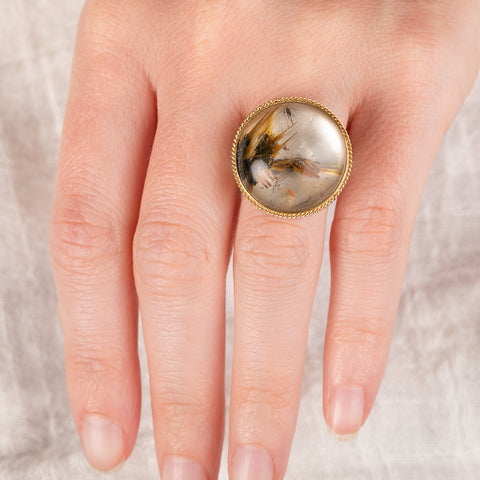 A hand wearing a ring with rutilated quartz set in an 18k gold bezel, with a sterling silver band