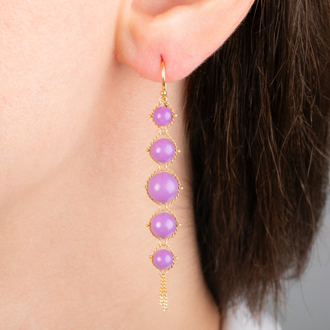A close-up of a person's ear wearing a long gold dangling earring with five round lavender-purple phosphosiderite stones.