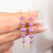 A close-up of a hand holding a pair of gold drop earrings with purple phosphosiderite stones, against a soft white background.