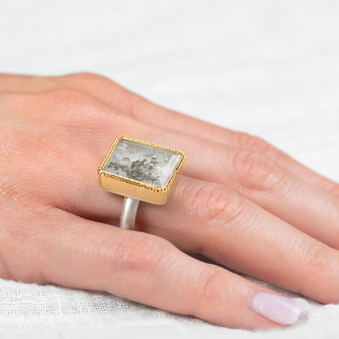 Gold and silver ring featuring a quartz with pyrite stone on a person's finger with a white background