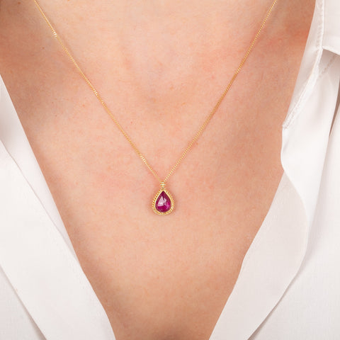 Gold necklace with a teardrop-shaped red ruby gemstone pendant on a person wearing a white shirt.