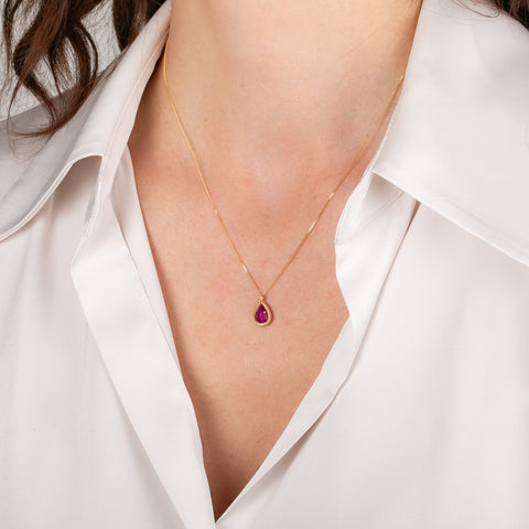 Gold necklace with a teardrop-shaped ruby pendant worn by a person in a white shirt.