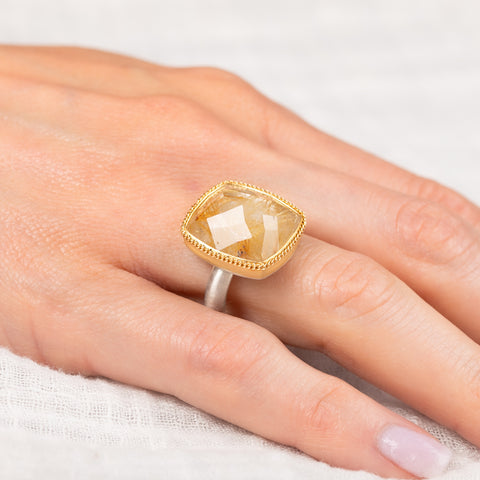 Gold and silver ring with a rutilated quartz gemstone on a person's finger against a neutral background