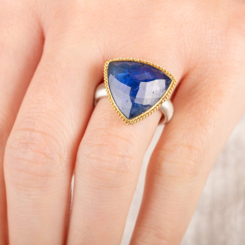 Close up of a person wearing a handmade ring with a tanzanite gemstone, gold bezel and sterling silver band on a white background