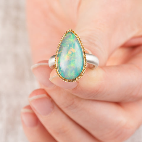 Close up of a hand holding an Ethiopian opal teardrop ring with gold bezel and silver band