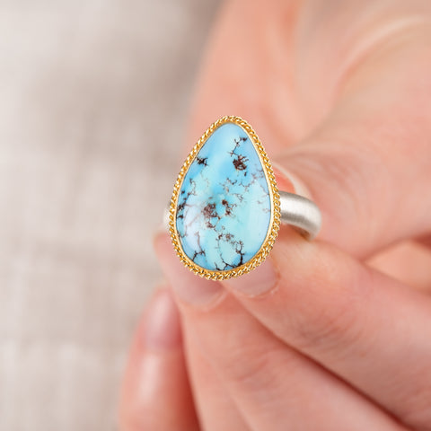 Close up of hand holding a turquosie teardrop ring with gold bezel and sterling silver ring