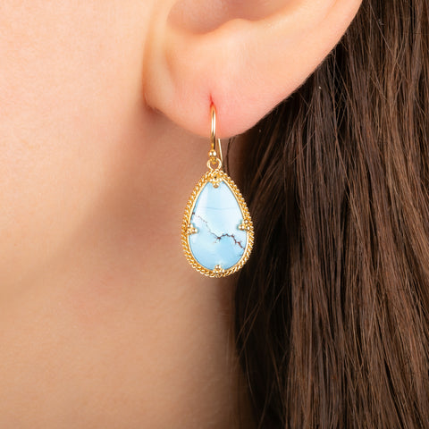 A close-up of a gold dangle earring with a light blue turquoise stone, worn on a person's ear.