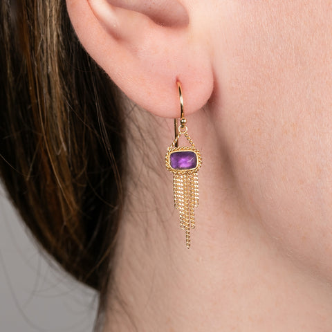 A person wears a handcrafted 18k gold earring featuring amethyst gemstones and a gold tassel adornment
