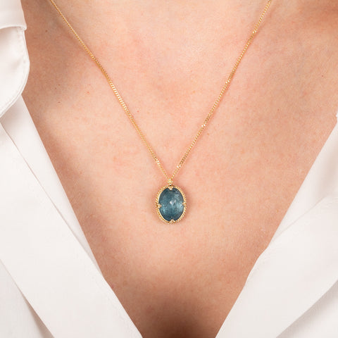 Gold necklace with an oval aquamarine pendant worn by a person in a white shirt
