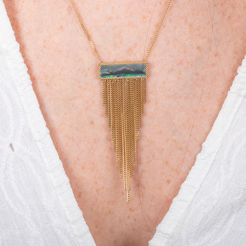 A person wears a handcrafted fine jewelry necklace featuring a black opal gemstone and 18k gold tassel adornment