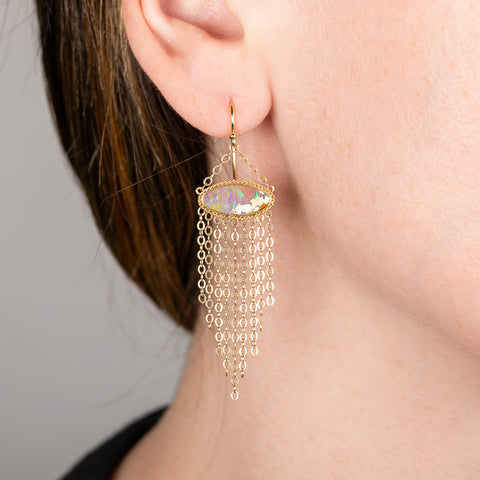 A person wears a handcrafted 18k gold earring featuring a one of a kind boulder opal gemstone and gold tassel adornment