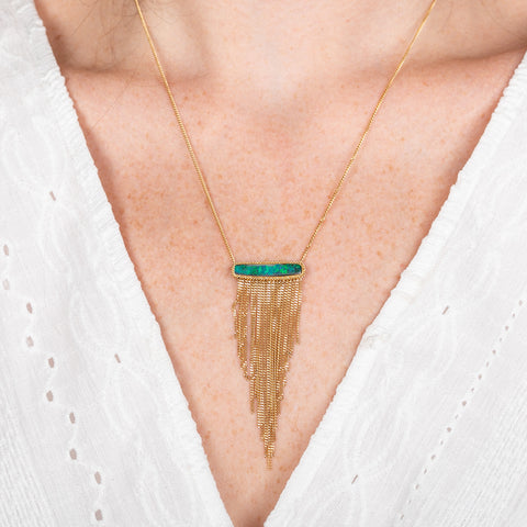 A person wears a handcrafted necklace featuring a boulder opal gemstone and 18k gold tassel adornment