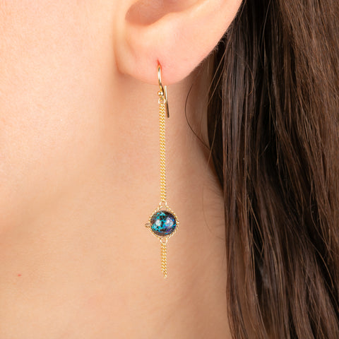 A close-up of a gold dangle earring worn on a person's ear, featuring a round iridescent boulder opal stone with a long gold chain