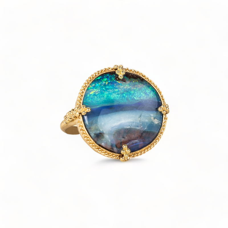 A rectangular opal with blue, purple and green hues is set in an 18k yellow gold bezel wrapped in chain with four beaded prongs on a thin ring band.