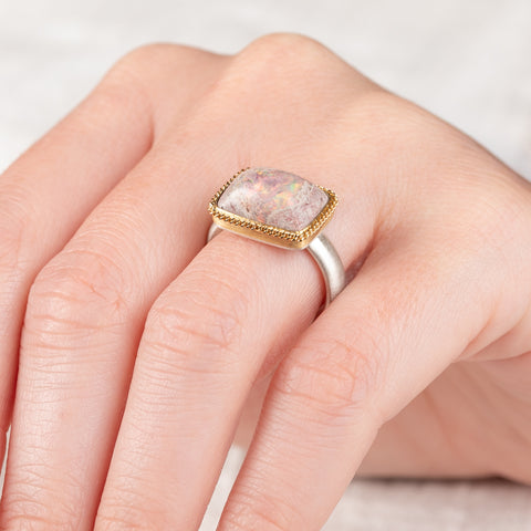 A person wears a handcrafed ring featuring a mexican opal gemstone in an 18k gold setting with a sterling silver band