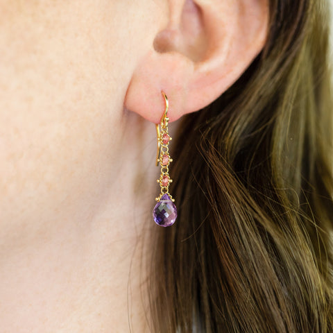A model wears an 18k yellow gold drop earring that features three woven spinel beads and a teardrop shaped amethyst hanging from a french hook closure