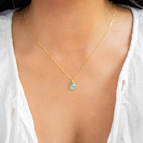 A model wears a teardrop shaped aquamarine stone necklace that is set in an 18k yellow gold bezel with braided details and granulated prongs. The pendant hangs on a thin gold chain.